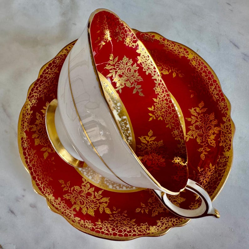 May include: A white teacup with a gold rim and handle sits on a red saucer. The saucer and the inside of the cup are red with a gold floral pattern. The edges of the saucer are scalloped and trimmed with gold.