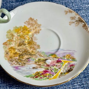 May include: A white porcelain saucer with a scalloped edge and a gold rim, featuring a colorful landscape illustration of a stone bridge over a stream, surrounded by trees and flowers, with a partial view of a matching teacup handle on the left.