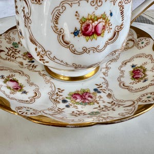 K&ouml;nnte beinhalten: Ein wei&szlig;es Tee-Set mit einer floralen Gestaltung. Die Tasse und Untertasse haben einen goldenen Rand und ein detailliertes Muster aus rosa Rosen und anderen Blumen. Das Set ist wahrscheinlich aus Porzellan und f&uuml;r den Teeservice bestimmt.