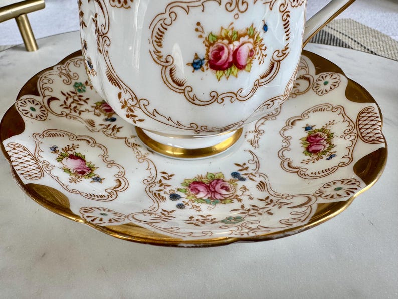 K&ouml;nnte beinhalten: Ein wei&szlig;es Tee-Set mit einer floralen Gestaltung. Die Tasse und Untertasse haben einen goldenen Rand und ein detailliertes Muster aus rosa Rosen und anderen Blumen. Das Set ist wahrscheinlich aus Porzellan und f&uuml;r den Teeservice bestimmt.