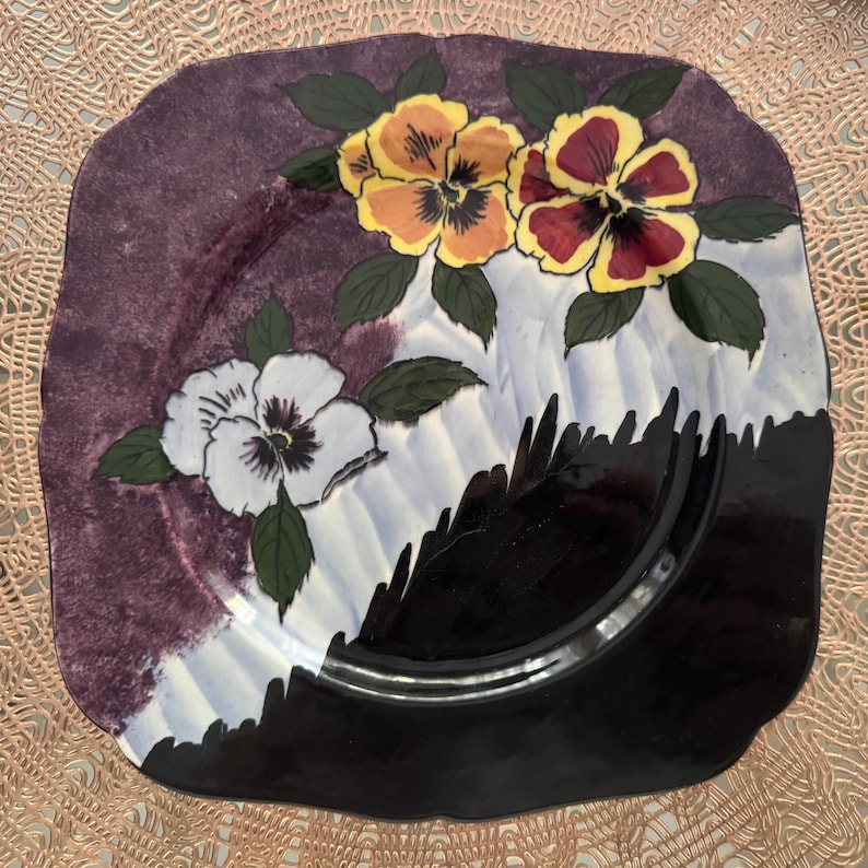 May include: Square ceramic plate with a floral design. The plate features a purple and black border with a white and light blue center. The plate is decorated with yellow, orange, white, and burgundy pansies and green leaves.