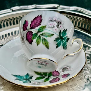 May include: White teacup and saucer set with gold trim. The teacup and saucer are decorated with a floral design featuring purple, green, and white leaves and flowers. The set is placed on a silver tray.