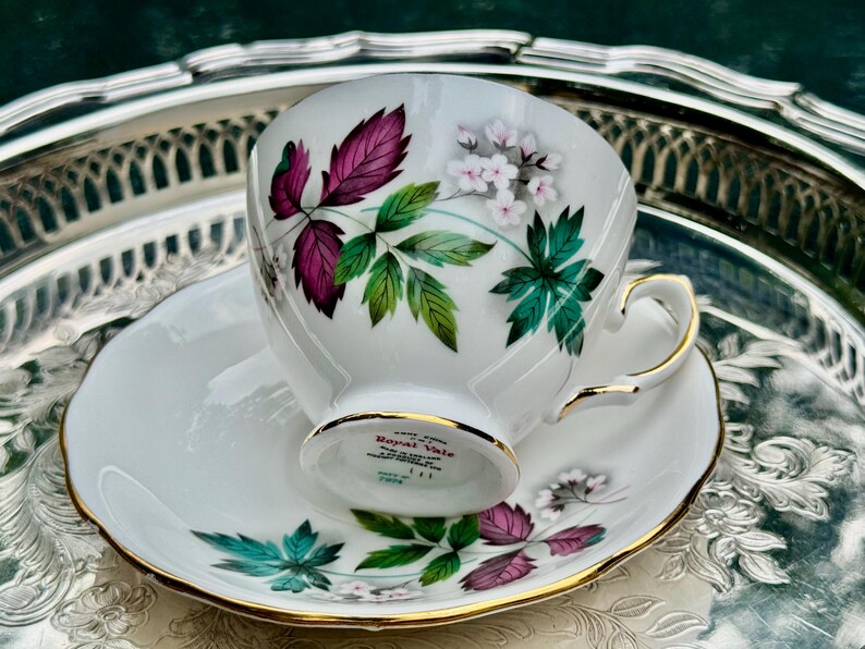 May include: White teacup and saucer set with gold trim. The teacup and saucer are decorated with a floral design featuring purple, green, and white leaves and flowers. The set is placed on a silver tray.