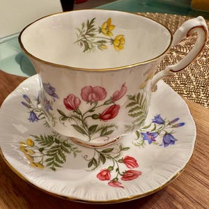 May include: A white teacup and saucer set with a floral pattern. The cup and saucer feature a delicate design of pink, red, blue, and yellow flowers on a white background.