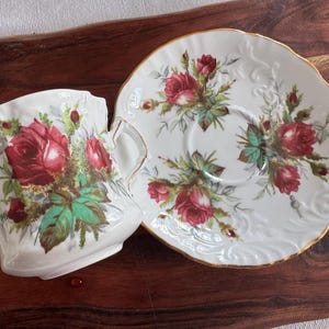 May include: A white teacup and saucer set with a floral design featuring red roses and green leaves. The set has a gold trim around the edges.