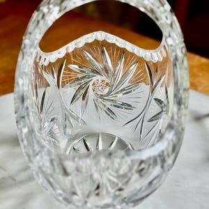 May include: A clear, cut-glass basket with a handle. The oval-shaped bowl features intricate starburst and linear patterns. The handle and rim are also detailed with cut glass designs, creating a sparkling effect.