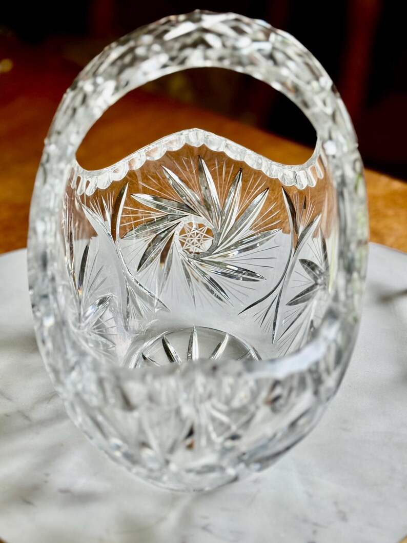 May include: A clear, cut-glass basket with a handle. The oval-shaped bowl features intricate starburst and linear patterns. The handle and rim are also detailed with cut glass designs, creating a sparkling effect.