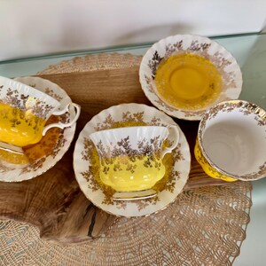 Puede incluir: Cuatro tazas y platillos de t&eacute; vintage. Las tazas y los platillos son blancos con ribetes dorados y un interior amarillo. Los platillos tienen un dise&ntilde;o floral. Las tazas de t&eacute; est&aacute;n sobre una superficie de madera.