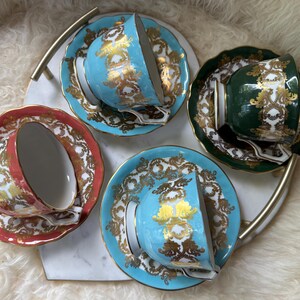 以下が含まれることがあります： ターコイズ、コーラル、グリーンを基調とした4つのヴィンテージティーカップとソーサー。それぞれに金色のフィリグリー模様が施されています。ティーカップは白と金のサービングトレイに置かれ、エレガントなティーセットを形成しています。