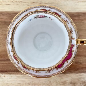 May include: Overhead view of a white teacup and saucer set with a floral design and gold trim. The cup and saucer have a magenta border and are set on a wooden surface. The teacup handle is gold.