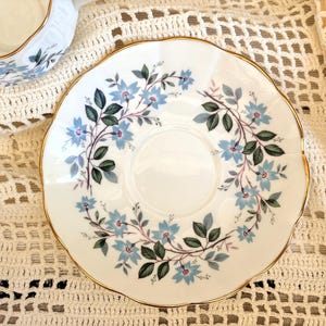 May include: A white porcelain saucer with a scalloped edge and a gold-trimmed rim. The center features a circular indentation surrounded by a delicate floral pattern of light blue flowers and dark green leaves arranged in a circular vine motif.