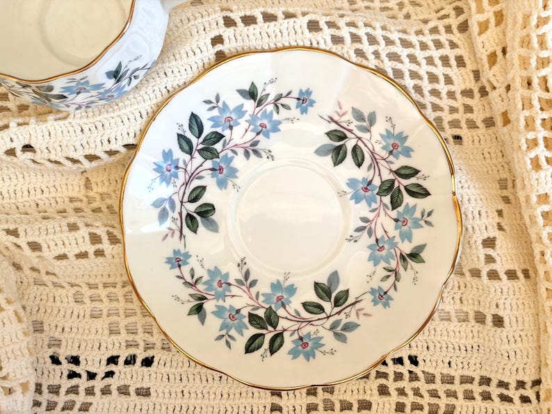 May include: A white porcelain saucer with a scalloped edge and a gold-trimmed rim. The center features a circular indentation surrounded by a delicate floral pattern of light blue flowers and dark green leaves arranged in a circular vine motif.