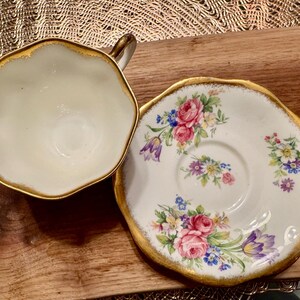 May include: A white teacup and saucer with gold trim and floral designs. The cup has a scalloped edge and the saucer has a floral pattern with pink, purple, and yellow flowers.