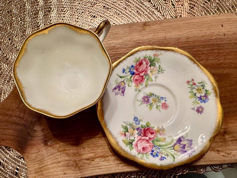 May include: A white teacup and saucer with gold trim and floral designs. The cup has a scalloped edge and the saucer has a floral pattern with pink, purple, and yellow flowers.