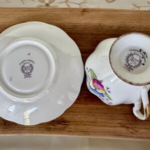 May include: White teacup and saucer set with floral and bird designs. The teacup has a gold handle and the saucer has a scalloped edge. The set is marked "Country Gardens Pansy Queen Anne Fine Bone China England."