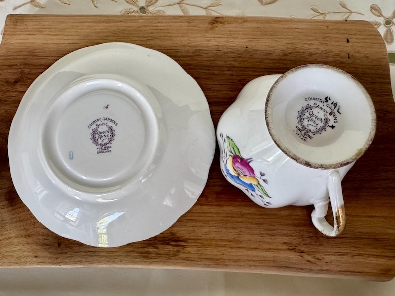 May include: White teacup and saucer set with floral and bird designs. The teacup has a gold handle and the saucer has a scalloped edge. The set is marked "Country Gardens Pansy Queen Anne Fine Bone China England."