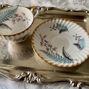 Puede incluir: Un juego de taza y platillo blancos con borde dorado y dise&ntilde;o de helechos y flores. La taza y el platillo est&aacute;n sobre una bandeja plateada con bordes ornamentados. La taza tiene asa.