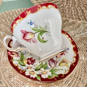 May include: A white teacup and saucer set with a red border and gold trim. The cup and saucer are decorated with floral designs in shades of red, pink, yellow, and blue. The set is designed for tea.