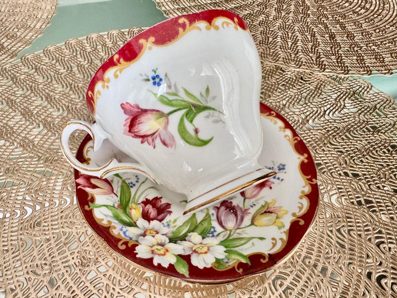 May include: A white teacup and saucer set with a red border and gold trim. The cup and saucer are decorated with floral designs in shades of red, pink, yellow, and blue. The set is designed for tea.
