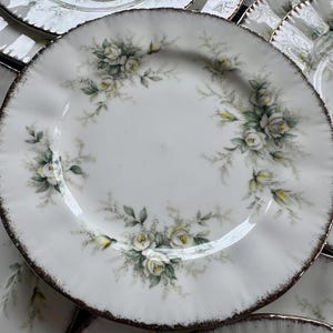 May include: A set of white ceramic plates with a delicate floral design featuring white roses and green leaves. The plates have a scalloped edge and a gold rim.