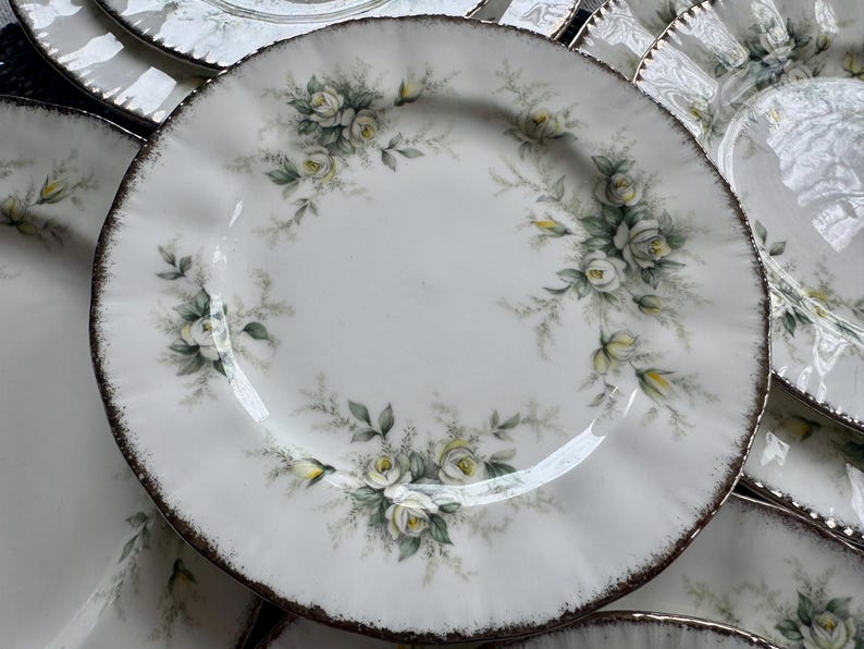 May include: A set of white ceramic plates with a delicate floral design featuring white roses and green leaves. The plates have a scalloped edge and a gold rim.