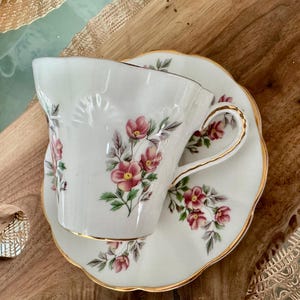 May include: A white teacup and saucer with a floral pattern. The cup has a gold rim and a delicate handle. The saucer has a scalloped edge and a gold rim.