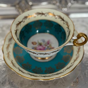 May include: A vintage teacup and saucer set. The teacup has a teal interior with gold accents and a floral design in the center. The saucer is teal with gold detailing. The set is cream-colored with gold trim.