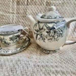 May include: A cream-colored teapot and matching teacup with saucer, both featuring a detailed black floral and botanical pattern. The teapot has a lid and a curved handle, while the teacup sits on a saucer with a matching rim design.