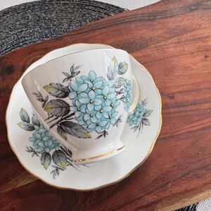 May include: A white teacup and saucer with a blue and green floral pattern. The cup and saucer have a gold trim.