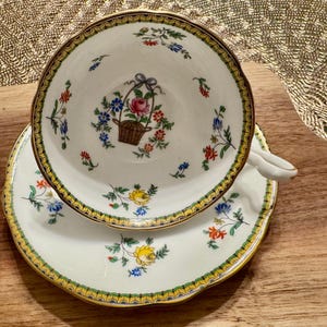 Pu&ograve; includere: Una tazza da t&egrave; e un piattino bianchi con un bordo dorato e un motivo floreale. La tazza presenta un cesto di fiori al centro, circondato da motivi floreali pi&ugrave; piccoli. Il piattino ha un motivo floreale simile.