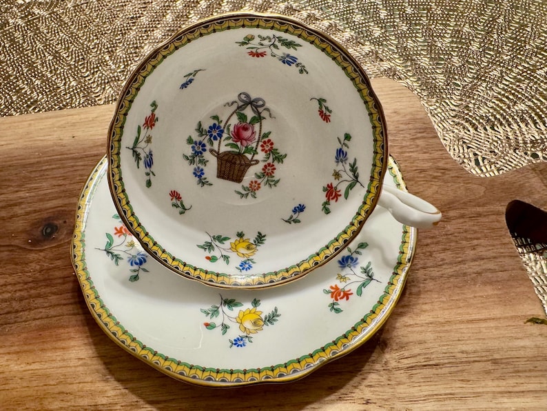 Pu&ograve; includere: Una tazza da t&egrave; e un piattino bianchi con un bordo dorato e un motivo floreale. La tazza presenta un cesto di fiori al centro, circondato da motivi floreali pi&ugrave; piccoli. Il piattino ha un motivo floreale simile.