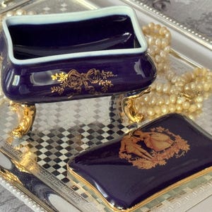 May include: A navy blue porcelain trinket box with a matching lid, featuring gold accents and a decorative design. The box sits on gold-colored feet and is displayed on a silver tray, with a pearl necklace in the background.