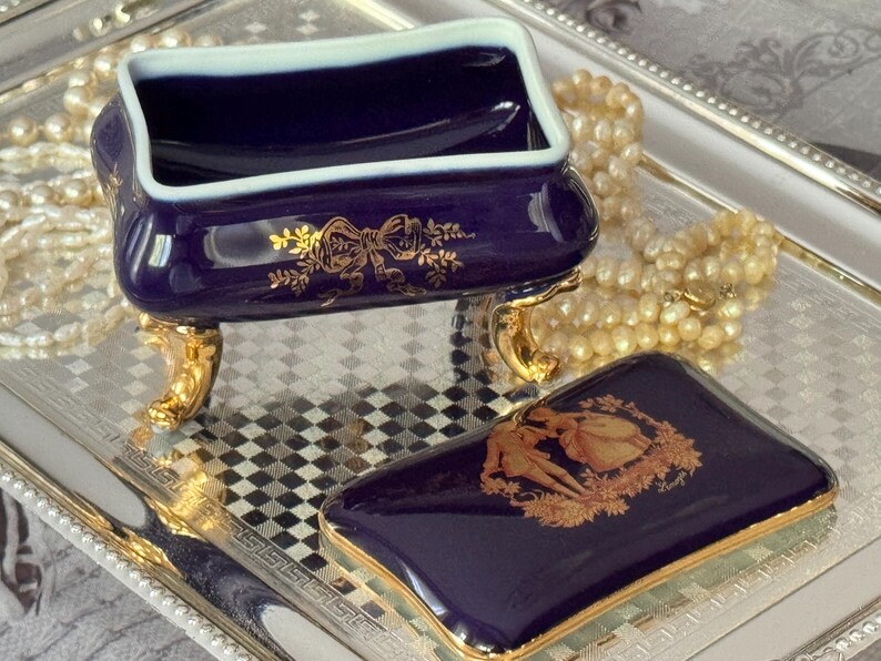 May include: A navy blue porcelain trinket box with a matching lid, featuring gold accents and a decorative design. The box sits on gold-colored feet and is displayed on a silver tray, with a pearl necklace in the background.