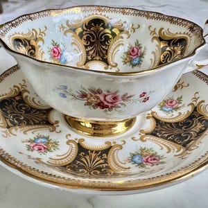 May include: A vintage teacup and saucer set. The white porcelain cup and saucer feature a black and gold ornate design with floral accents in pink, blue, and yellow. The rim and handle are trimmed in gold.