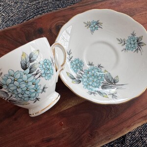 May include: A white teacup and saucer set with a blue and gray floral pattern. The floral design features delicate blue flowers and gray leaves.