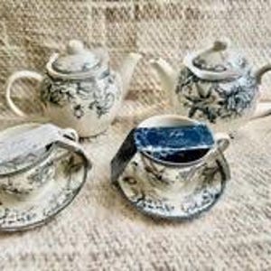 May include: A tea set consisting of two ceramic teapots and two matching teacups with saucers. Each piece features a white base with a detailed dark gray floral and botanical pattern. The teacups have dark rectangular labels attached to the rims.