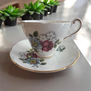 May include: A white teacup and saucer with a floral design featuring red, blue, and white flowers. The saucer has a gold trim.