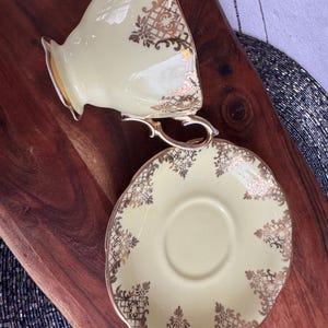 May include: A pale yellow teacup and saucer set with a gold floral pattern. The cup has a handle and the saucer has a scalloped edge.