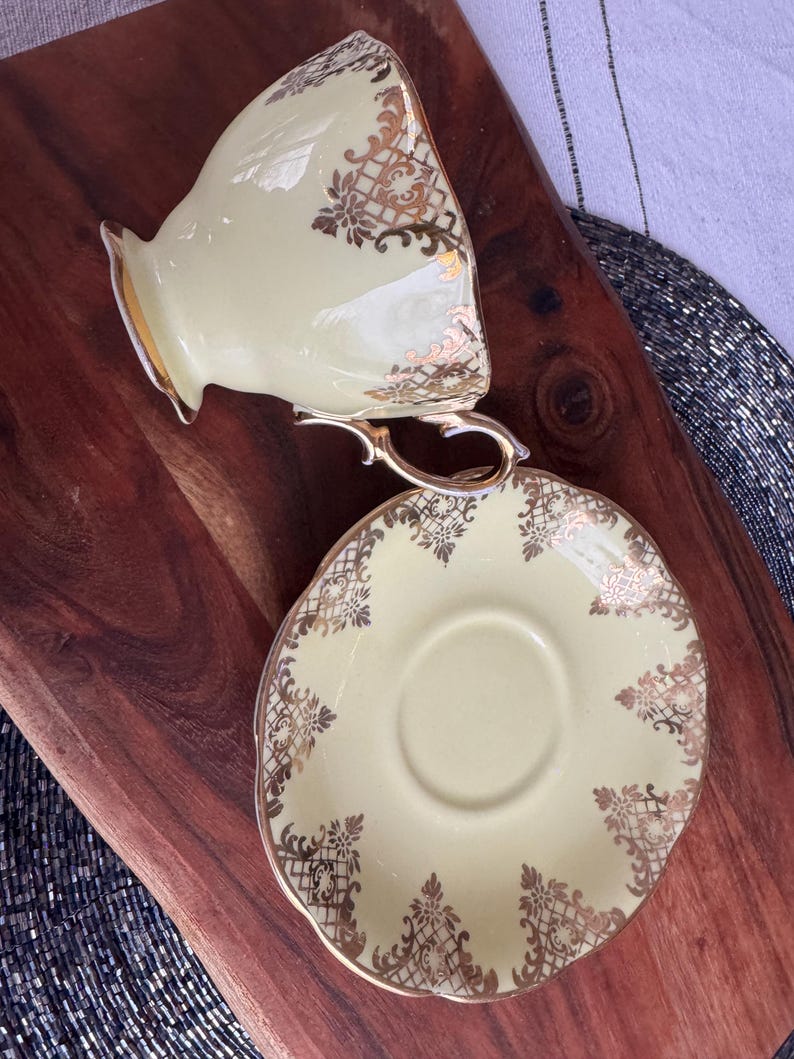May include: A pale yellow teacup and saucer set with a gold floral pattern. The cup has a handle and the saucer has a scalloped edge.