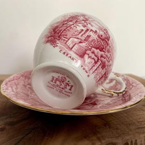 Pu&ograve; includere: Un set di tazza e piattino bianchi con un'illustrazione rossa di un edificio e la scritta "CASAR'S TOWER". La tazza e il piattino hanno un motivo floreale rosa e finiture dorate. La tazza &egrave; contrassegnata "Sutherland China Made in England".