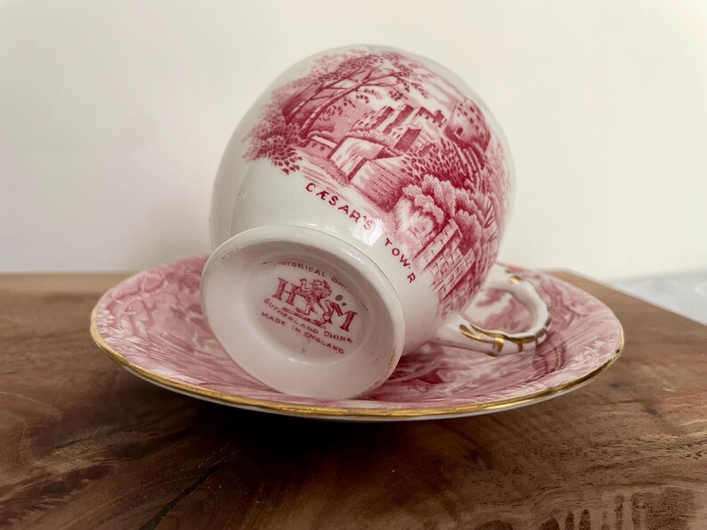 Pu&ograve; includere: Un set di tazza e piattino bianchi con un'illustrazione rossa di un edificio e la scritta "CASAR'S TOWER". La tazza e il piattino hanno un motivo floreale rosa e finiture dorate. La tazza &egrave; contrassegnata "Sutherland China Made in England".