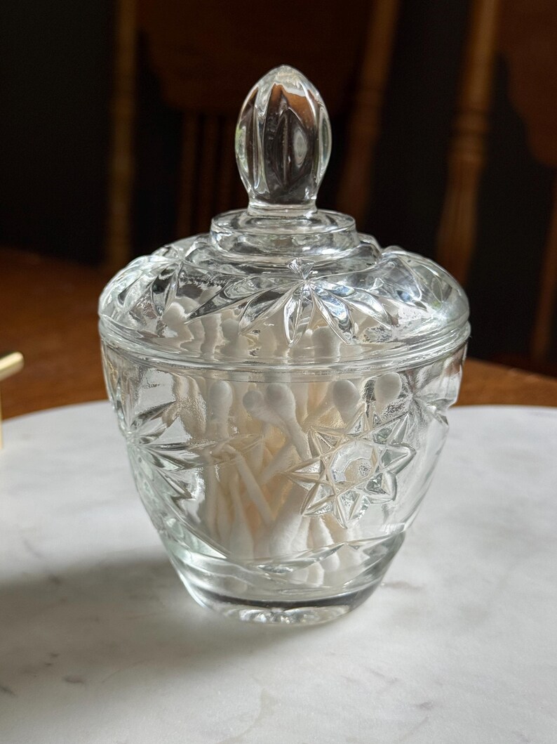 May include: A clear glass lidded jar featuring a pressed starburst pattern on the exterior. The container is filled with white cotton swabs and topped with a matching glass lid that has a pointed finial handle at the center of the lid.