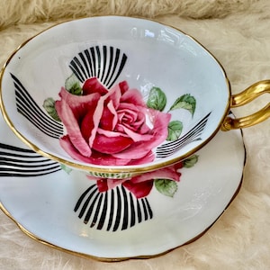 May include: White teacup and saucer set with a gold rim and handle. The teacup and saucer feature a large pink rose design with green leaves and black and white striped accents. The teacup is sitting on the saucer.