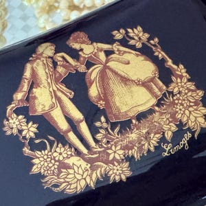 May include: A dark blue decorative box with a gold illustration of a couple in period clothing, surrounded by flowers. The word "Limoges" is written in gold script. The box has a glossy finish.