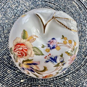 K&ouml;nnte beinhalten: Eine wei&szlig;e Porzellan-Schmuckschatulle in Apfelform mit einem goldenen Blattdesign und floralen Mustern in Rot-, Gelb-, Blau- und Gr&uuml;nt&ouml;nen.