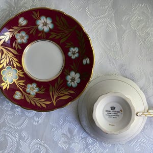 May include: A Royal Stafford bone china teacup and saucer set. The saucer features a deep burgundy rim with gold leaf accents and light blue flowers. The teacup is inverted, showing a white base with the Royal Stafford Made in England stamp.