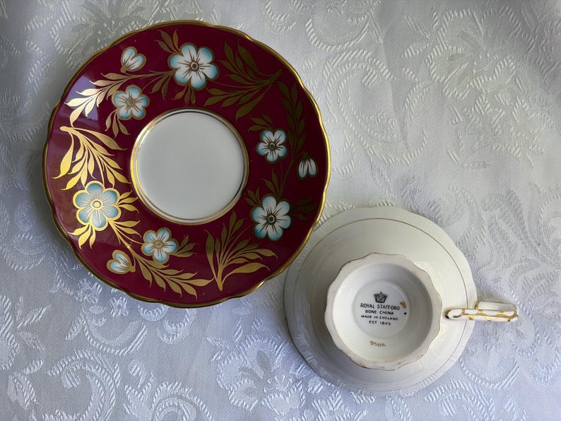 May include: A Royal Stafford bone china teacup and saucer set. The saucer features a deep burgundy rim with gold leaf accents and light blue flowers. The teacup is inverted, showing a white base with the Royal Stafford Made in England stamp.