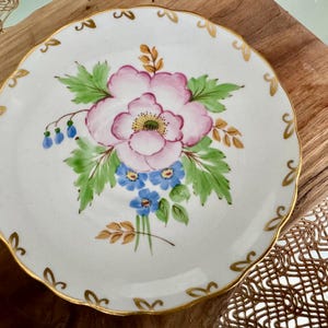May include: A white porcelain saucer with a gold rim and a floral design. The design features a large pink flower with blue and green accents.