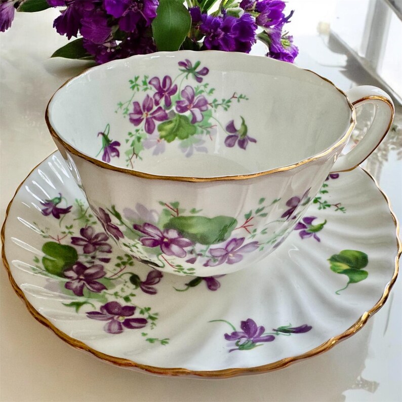K&ouml;nnte beinhalten: Wei&szlig;es Tee-Set mit einem Veilchen-Blumendesign. Tasse und Untertasse haben einen Goldrand. Die Tasse hat einen Henkel. Die Untertasse hat einen gewellten Rand. Das Set ist mit lila und gr&uuml;nen Blumenmustern verziert.