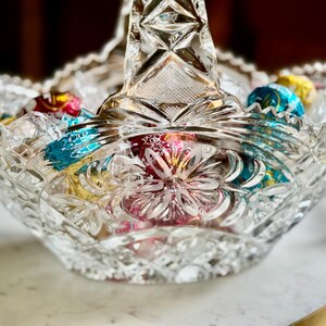 May include: A clear, cut-glass basket with a handle, filled with colorful foil-wrapped candies. The basket has a scalloped rim and a floral design on the base. The candies are in shades of red, blue, and yellow.